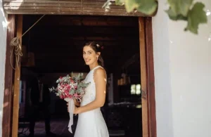 Novia sonriente con vestido blanco y ramo de flores rosas, posando en la entrada de un lugar rústico durante su boda en Valencia.